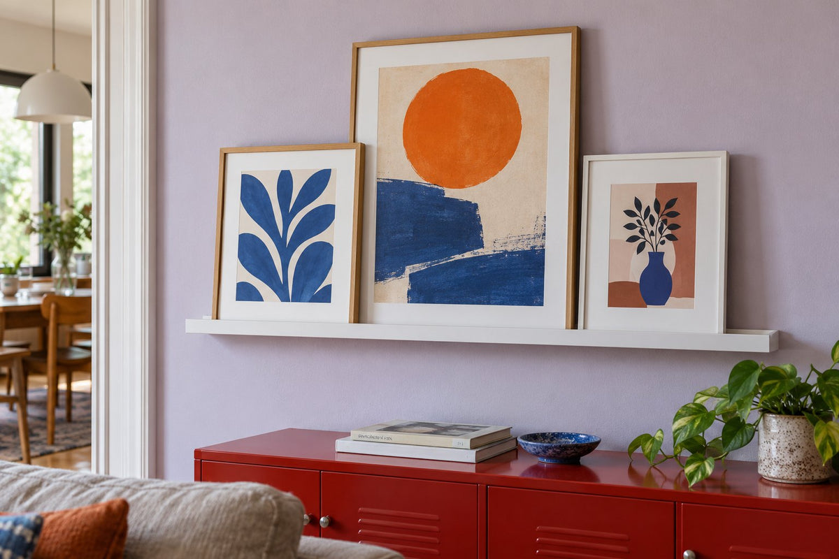 Trois cadres de tailles différentes sur une étagère blanche, mur lilas et meuble rouge.