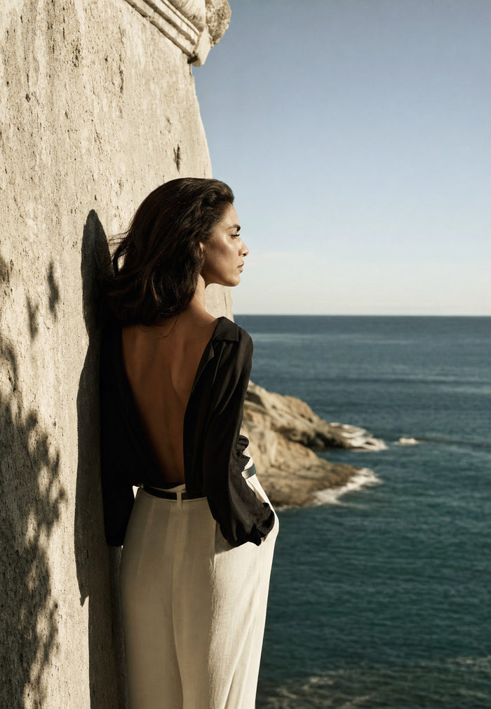 Adossée à un mur de pierre chaude, une femme en haut noir et pantalon crème regarde la mer, le profil tourné vers le large.