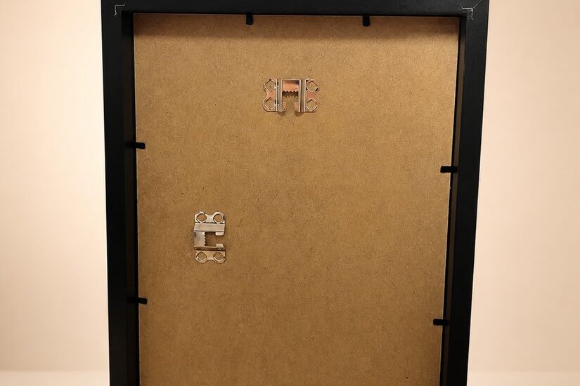 Dos d'un cadre noir : panneau de bois, attaches métalliques rabattables et deux pattes d'accrochage, l'une pour le portrait, l'autre pour le paysage.